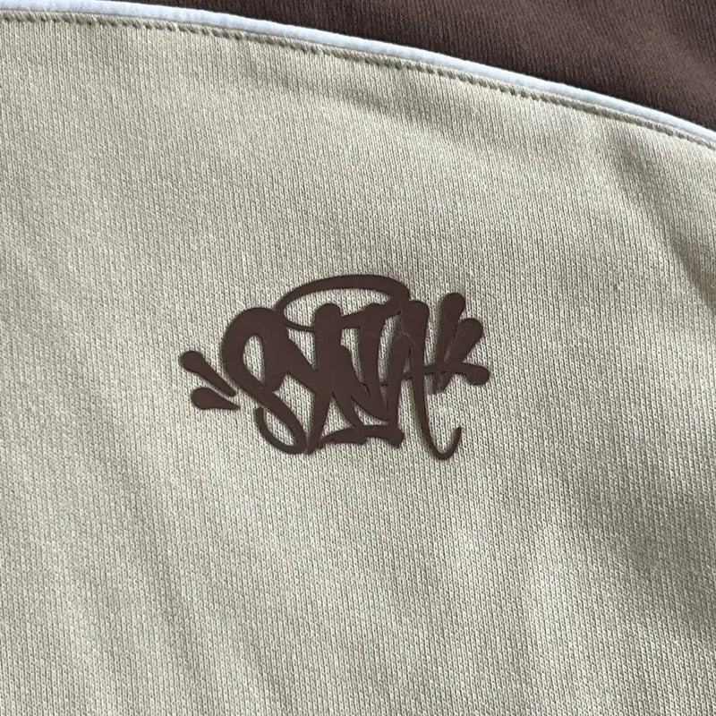 A close-up of the Syna World Brown Beige Two-Tone Letter Print Hoodie, showing beige fabric with a brown, graffiti-style monogram logo, plus brown material and white piping at the top.