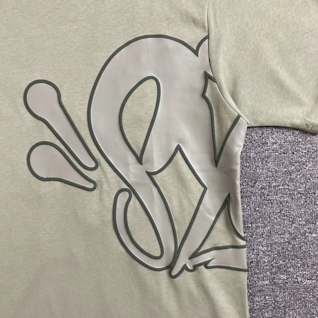 A close-up of the Syna World Green Letter Graphic Crew Neck T-Shirt displays its large abstract black design on light beige fabric, with the sleeve and part of the torso shown, lying on a textured gray surface.