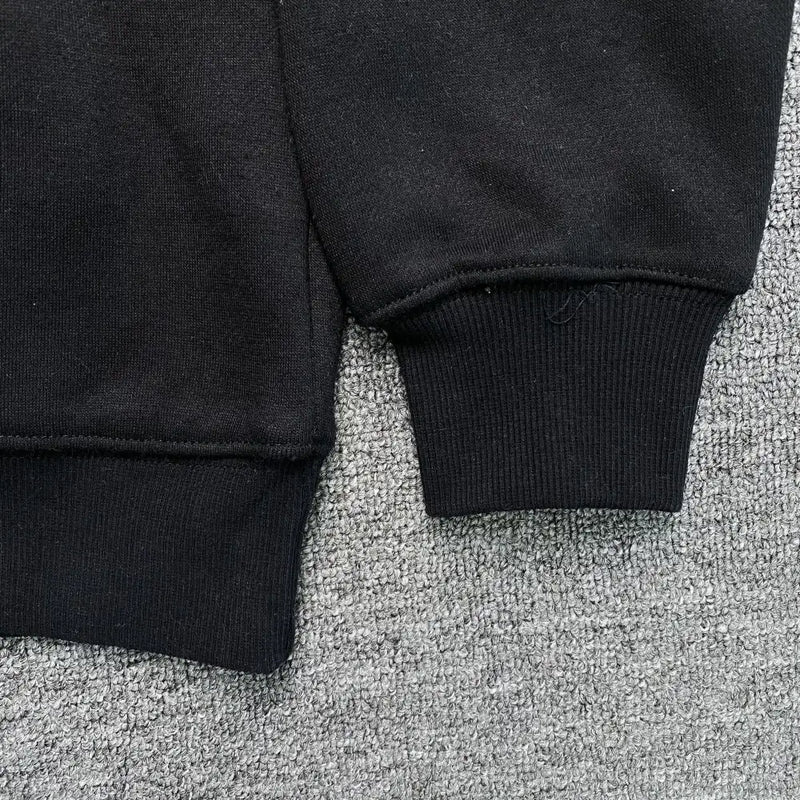 Close-up of the cuff and hem of the Syna World Black Yellow Graffiti Graphic Oversized Hoodie, laid flat on a gray surface. Soft fabric and visible stitching emphasize the premium streetwear quality.