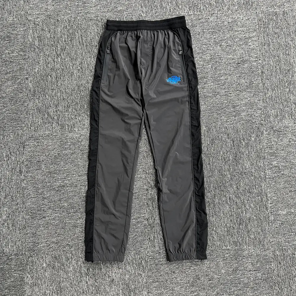 Syna World grey and black patchwork track pants with blue graffiti logo on left thigh, flat lay view