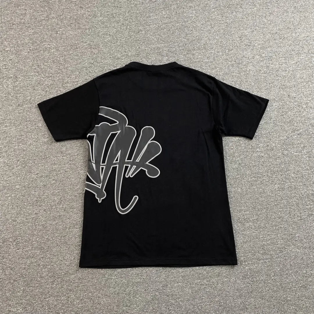 The Syna World Black Front Graphic Print T-Shirt, made from cotton, is displayed flat on a grey carpet with bold, stylized white graffiti text and design across the back—making a striking statement in streetwear fashion.