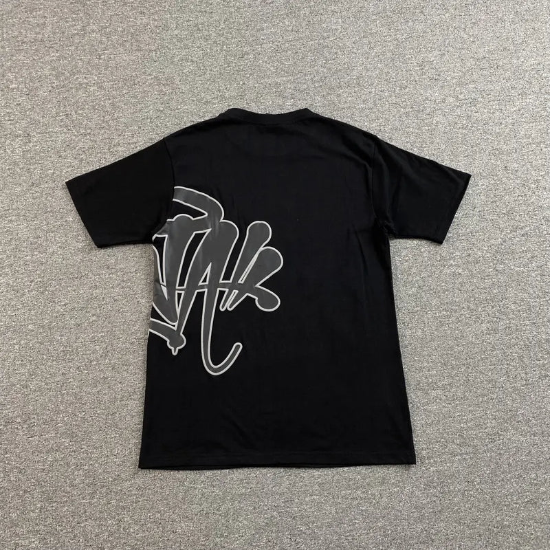 The Syna World Black Front Graphic Print T-Shirt, made from cotton, is displayed flat on a grey carpet with bold, stylized white graffiti text and design across the back—making a striking statement in streetwear fashion.