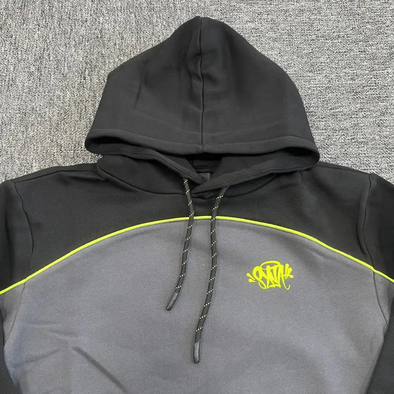 The Syna World Black Colorblock Neon Trim Pullover Hoodie, featuring neon yellow piping and a graffiti-style chest logo, is displayed flat on a textured gray carpet.