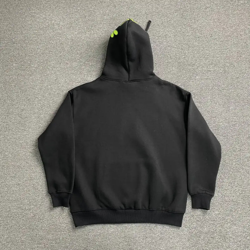 The Syna World Black Neon Green Graffiti Hood Pullover Hoodie is laid flat on a gray carpet, shown from the back, and features long sleeves, ribbed cuffs and hem, and two small neon green dots at the top of the hood.