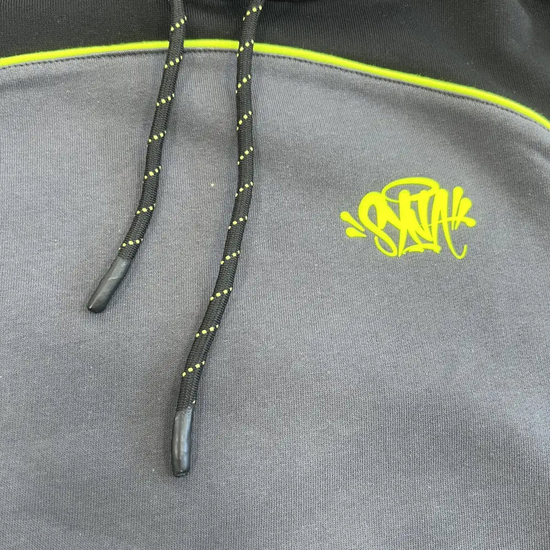 Close-up of the Syna World Black Colorblock Neon Trim Pullover Hoodie with black drawstrings featuring yellow dots, a neon yellow graffiti-style logo on the chest, and a neon yellow stripe across the shoulders.