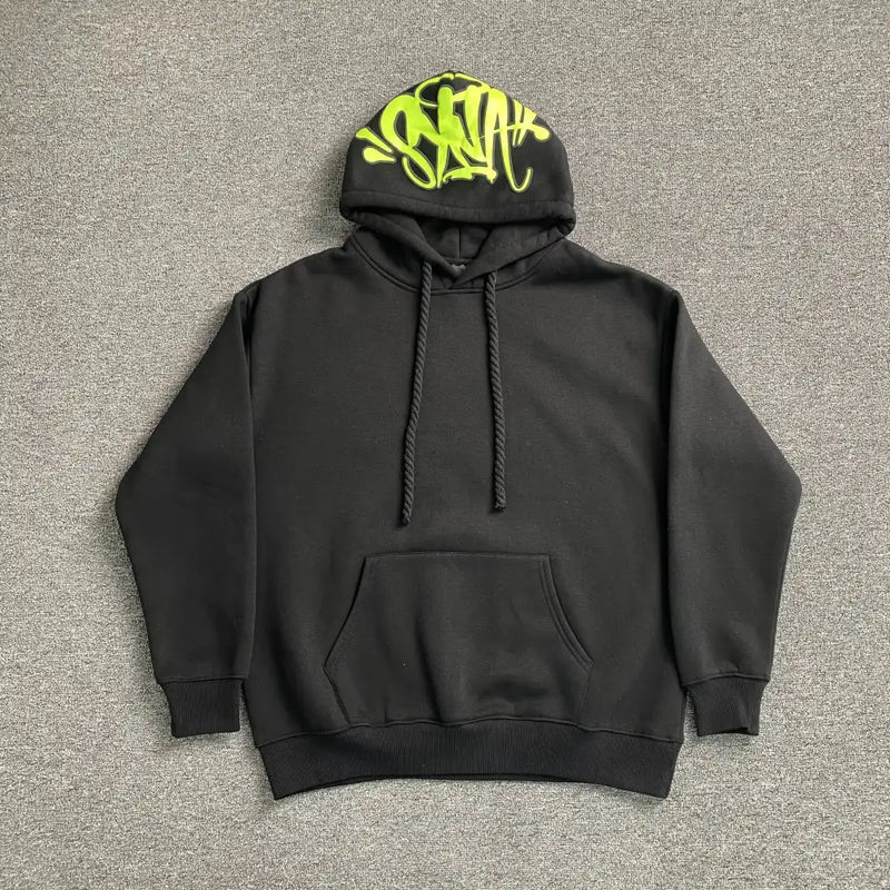 The Syna World Black Neon Green Graffiti Hood Pullover Hoodie features a front pocket and neon green graffiti-style text on the hood, making it a bold streetwear essential—shown laid flat on a gray textured surface.