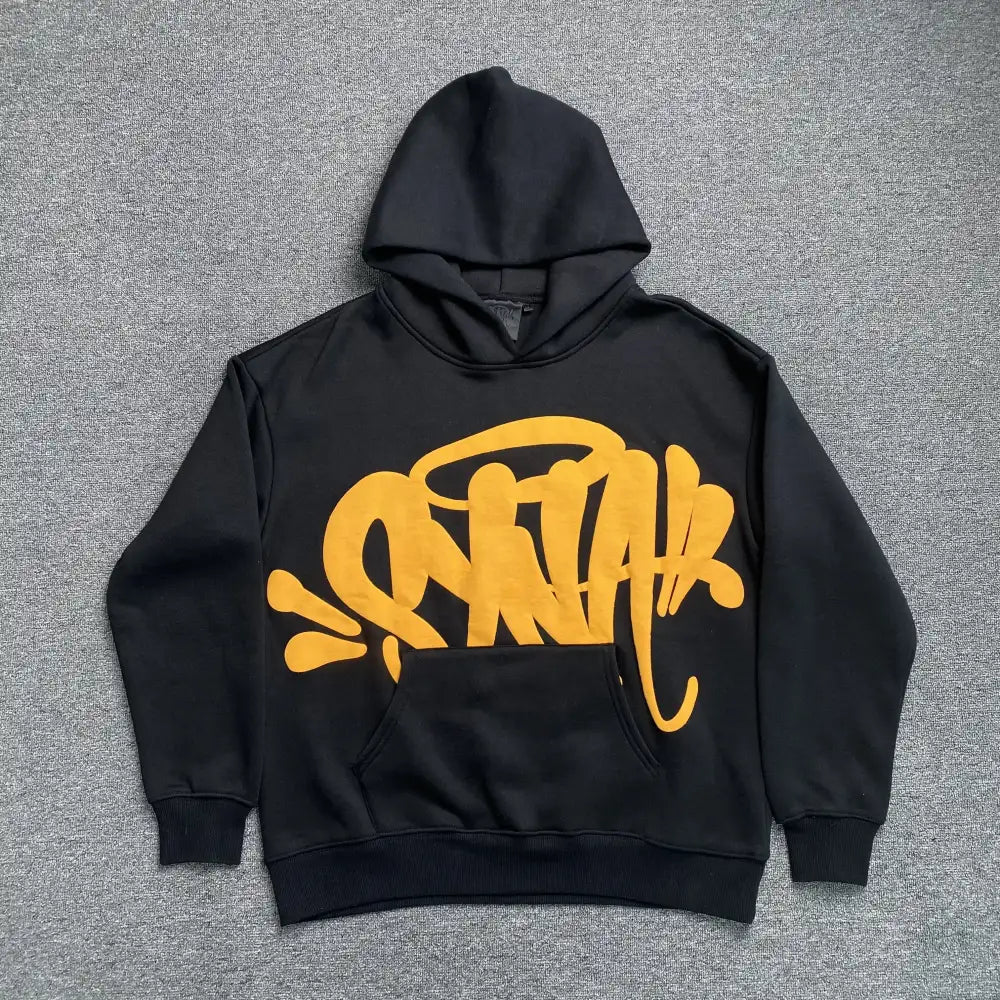 Black oversized hoodie with large yellow graffiti-style front graphic print, laid flat on grey surface