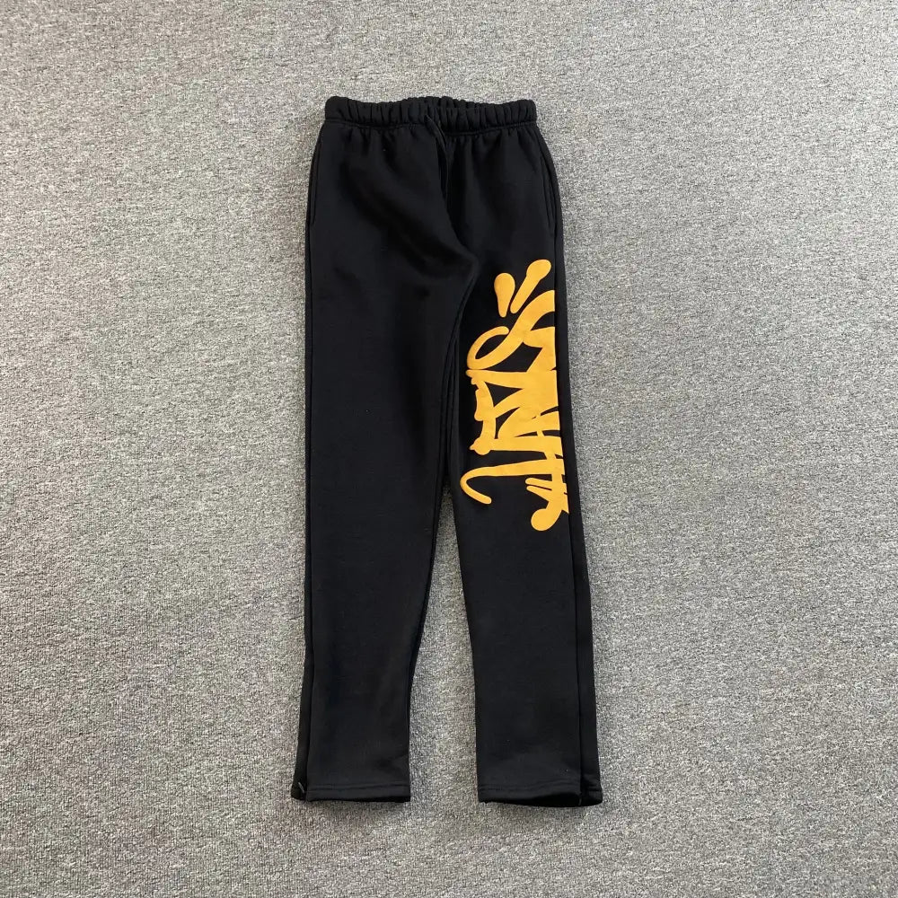 Black straight-leg jogger pants with yellow graffiti letter print on the left leg, laid flat on a grey surface