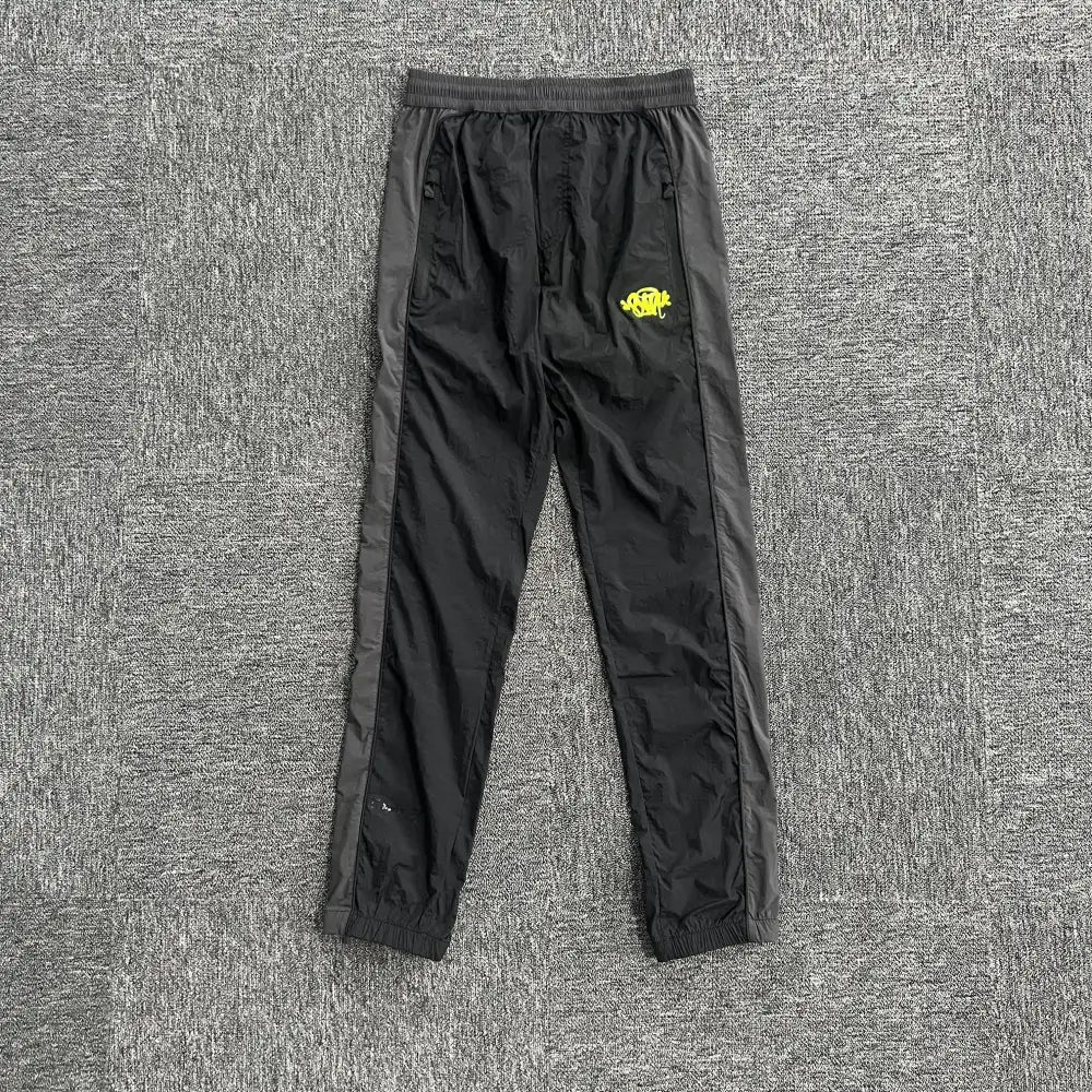 Syna World black track pants with yellow logo print and grey side panels laid flat