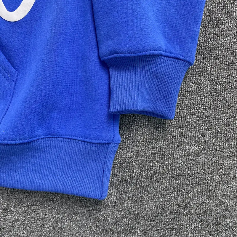 Close-up of the sleeve and waistband of the Syna World Blue Graffiti Letter Print Hoodie, featuring a white design on royal blue—laid on a textured gray surface, it’s a bold streetwear pullover for standout style.