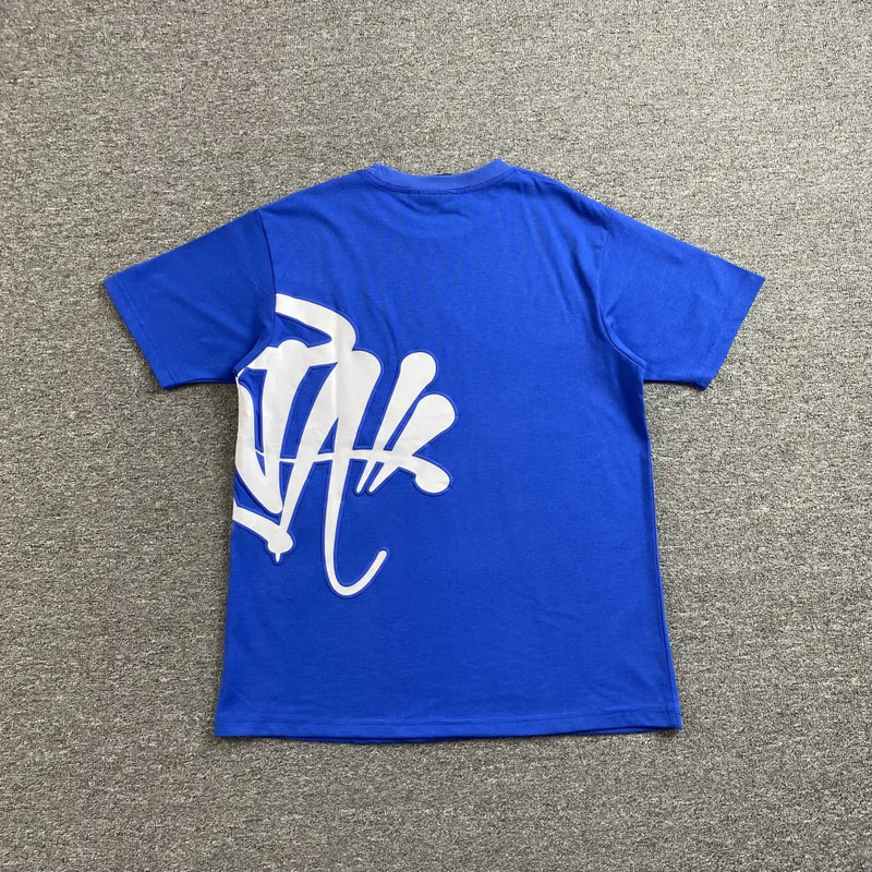 The Syna World Blue White Front Graphic Crew Neck T-Shirt lies flat on a gray carpet, featuring a bold white graffiti-style graphic on the lower left back—an essential statement piece for streetwear fans.