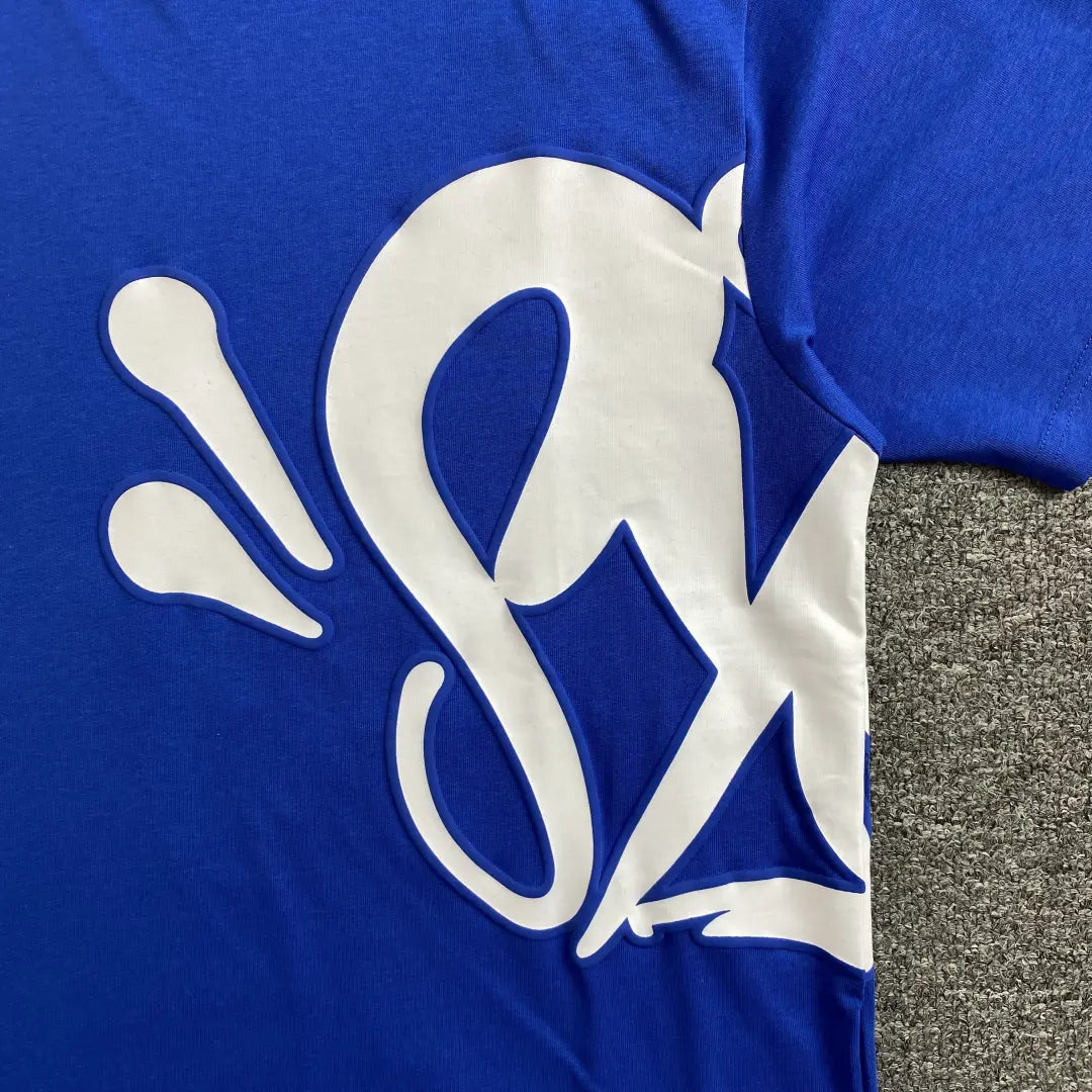Close-up of the Syna World Blue White Front Graphic Crew Neck T-Shirt, a cotton streetwear piece with a bold white stylized "S" and splash designs on blue fabric, displayed on a textured gray surface.