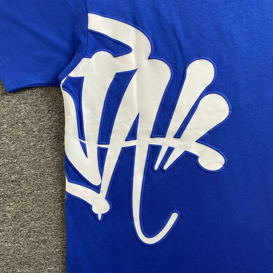 The Syna World Blue White Front Graphic Crew Neck T-Shirt features a bold white graffiti-style print across the front, shown laid flat on a textured gray surface that enhances its modern streetwear vibe.
