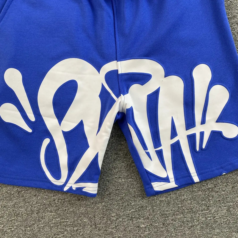 Syna World Blue Graffiti Logo Print Sweat Shorts featuring large white graffiti-style lettering and splash designs on the front, shown on a gray textured surface.