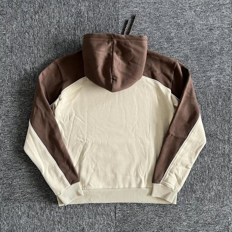 The Syna World Brown Beige Two-Tone Letter Print Hoodie features a beige body and inner sleeves with brown hood, shoulders, and outer sleeves for a modern contrast, shown flat on a gray textured surface.