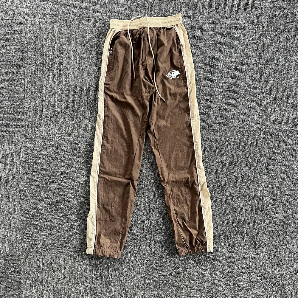Syna World brown jogger track pants with beige side stripes and logo print on left thigh, laid flat