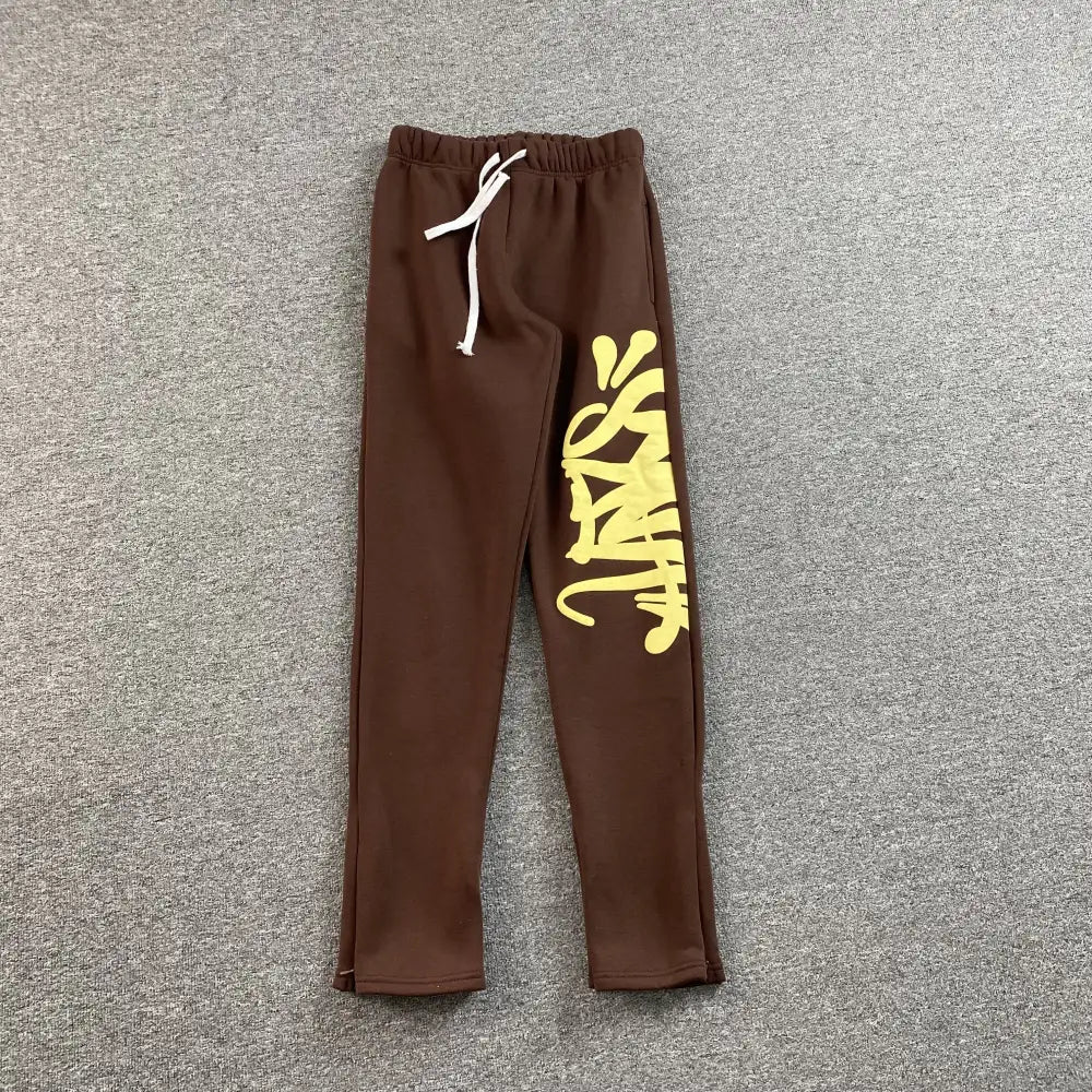 Brown Syna World sweatpants with yellow graffiti letter print on the left leg, laid flat