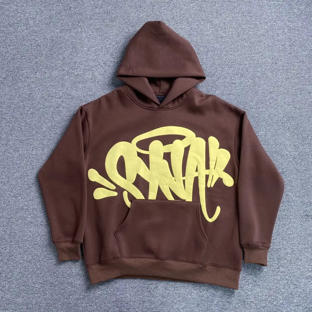Brown oversized hoodie with yellow graffiti letter print on the front, laid flat on a grey surface