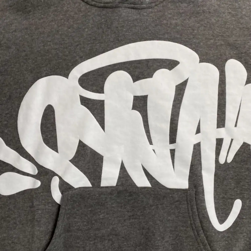 The Syna World Grey Graffiti Graphic Pullover Hoodie is a streetwear staple, showcasing a bold white graffiti design with dynamic shapes and lines across the chest, partially obscured by the front pocket.