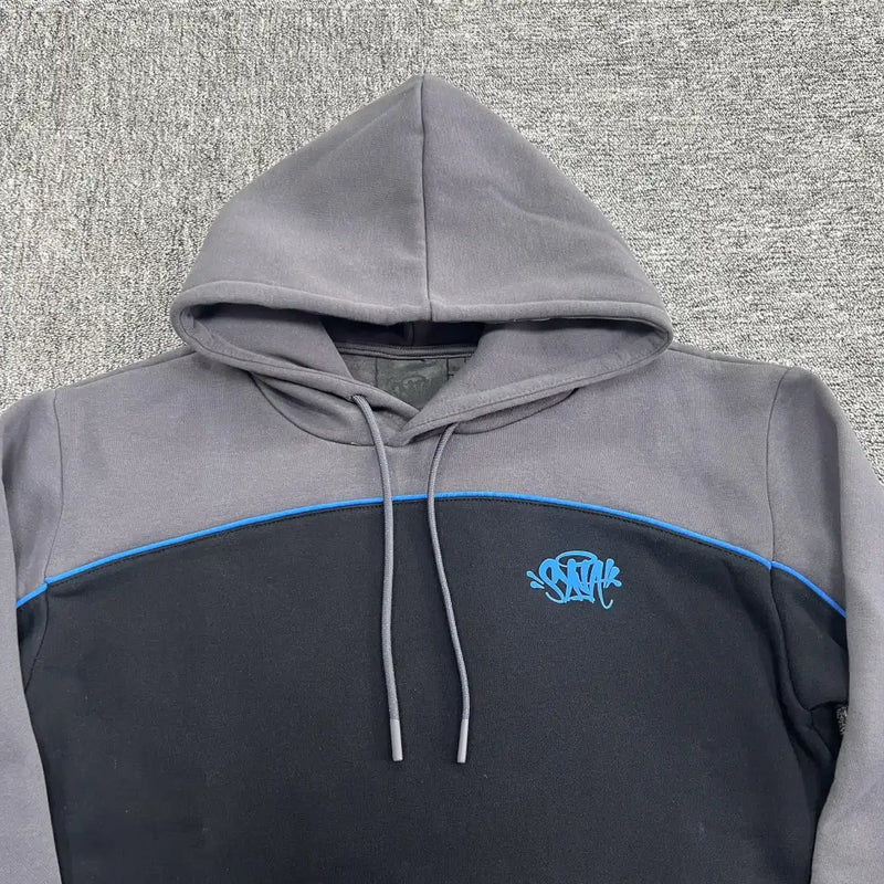 The Syna World Black Grey Color Block Graphic Hoodie features a gray hood and shoulders, black body, blue piping, and a blue embroidered chest design, shown on a gray textured surface.