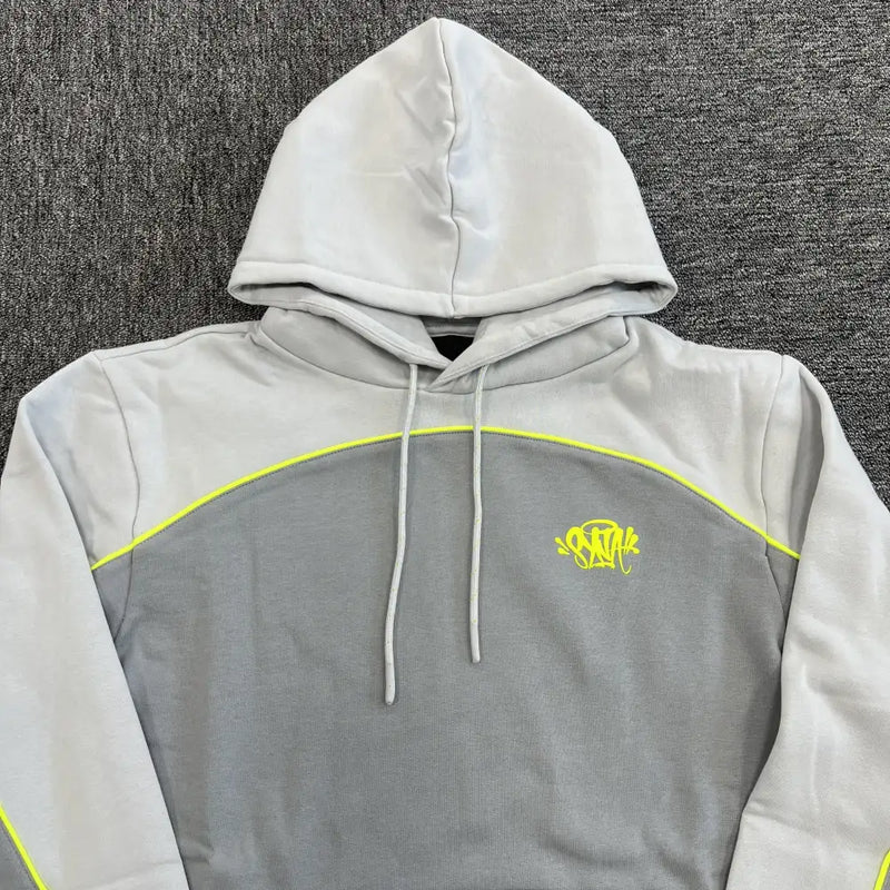 The Syna World Grey Colorblock Neon Trim Hoodie features a two-tone grey design with neon yellow piping, a hood with drawstrings, and a graffiti-style logo on the left chest, shown laid flat on a dark textured surface.