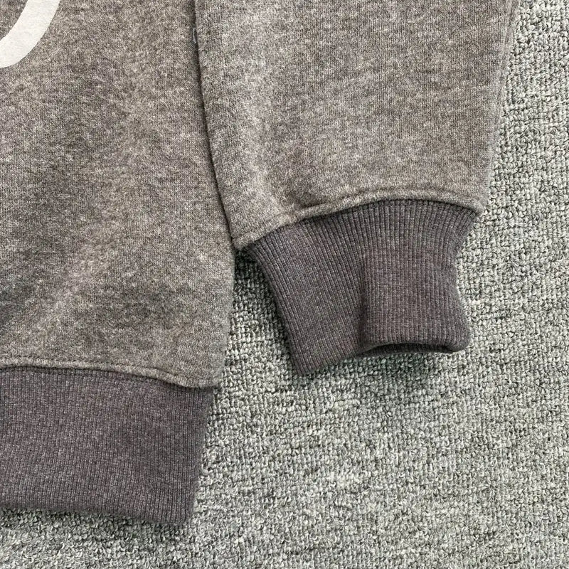 Close-up of the sleeve and hem of the Syna World Grey Graffiti Graphic Pullover Hoodie with ribbed cuffs, displayed on a textured gray carpet.