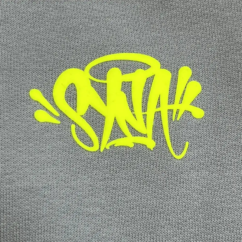 Yellow graffiti-style text pops against the gray fabric of the Syna World Grey Colorblock Neon Trim Hoodie, giving this streetwear piece an urban edge and artistic flair with its bold neon accents.