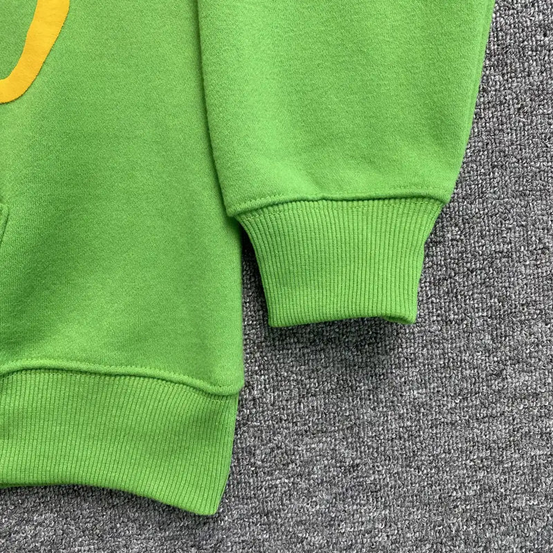 A close-up of the Syna World Green Graffiti Graphic Pullover Hoodie shows ribbed sleeves and waistband on a textured gray carpet, with a partial yellow graffiti front graphic adding streetwear flair to the left side.