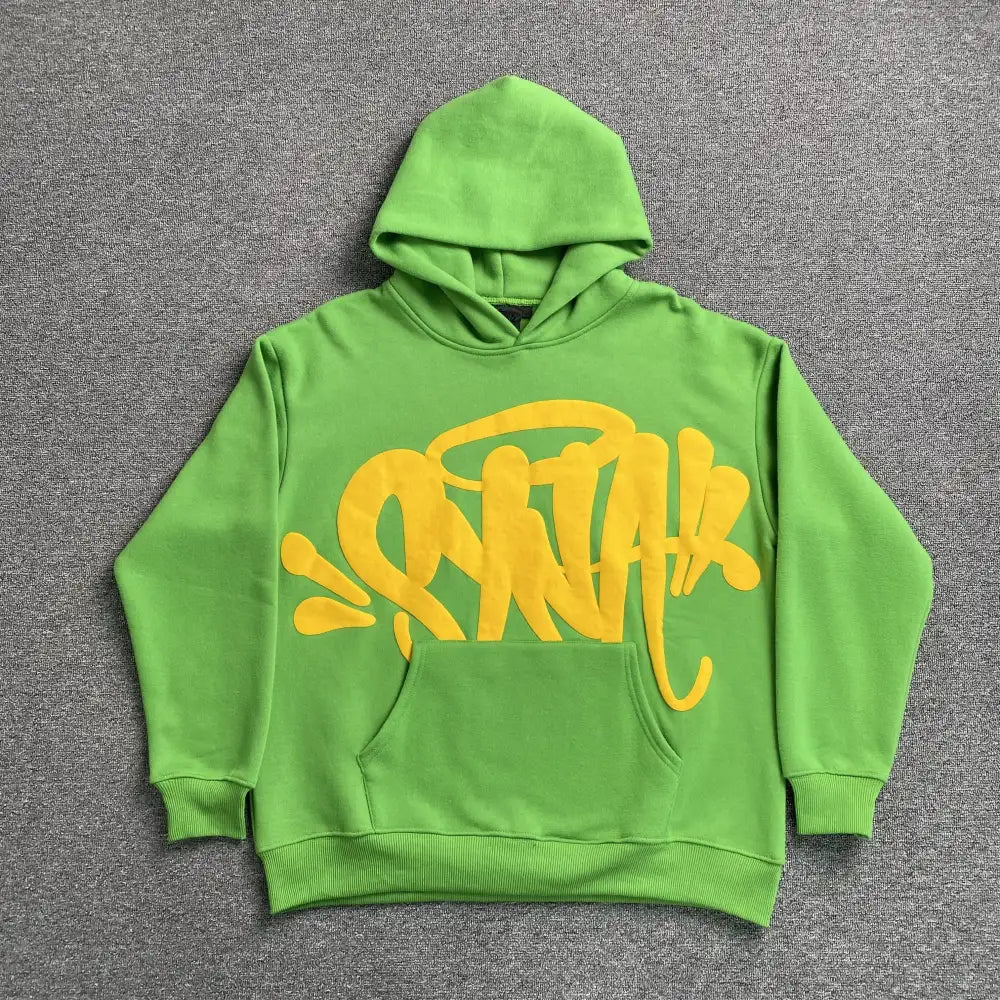Green pullover hoodie with yellow graffiti graphic on the front chest, laid flat on a grey surface