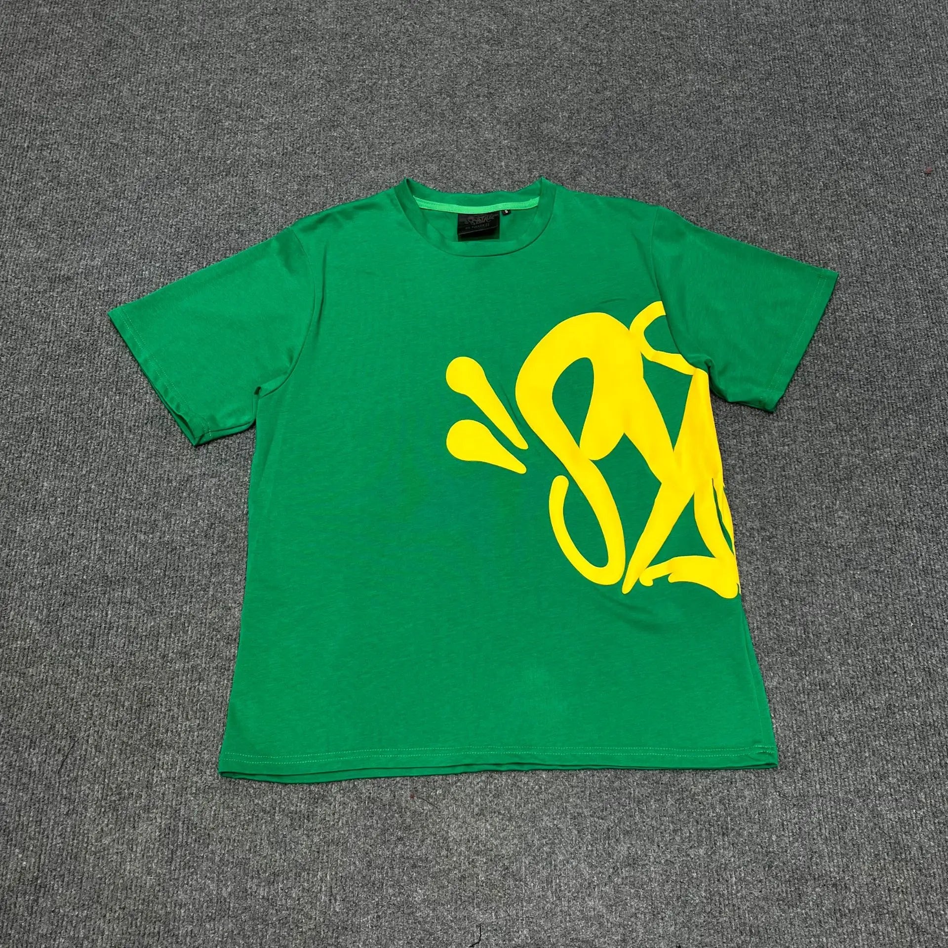 Green Syna World T-shirt with oversized yellow graffiti graphic print on the front, laid flat on grey surface