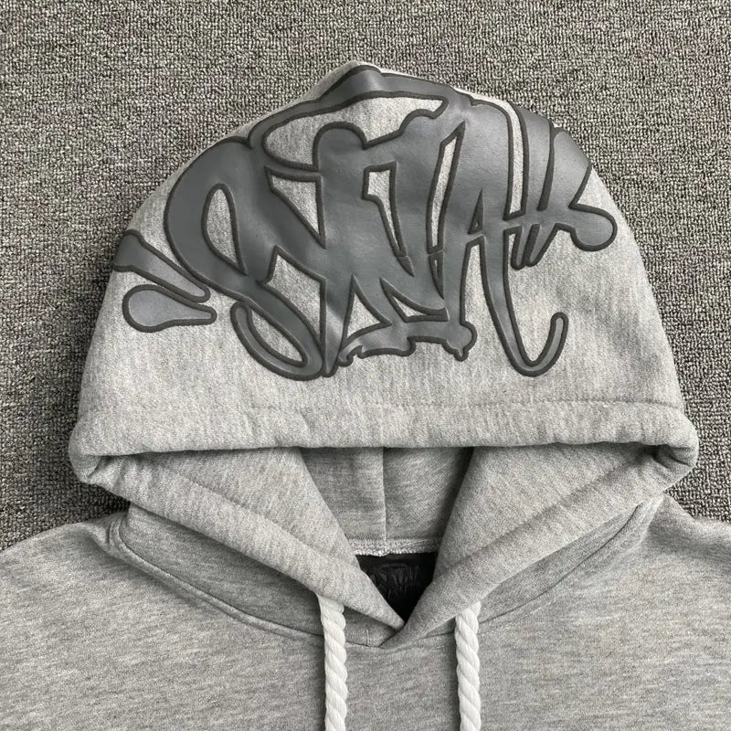 The Syna World Grey Graffiti Print Oversized Hoodie features thick white drawstrings and bold black graffiti lettering on the hood, displayed on a textured gray surface—ideal for standout streetwear style.