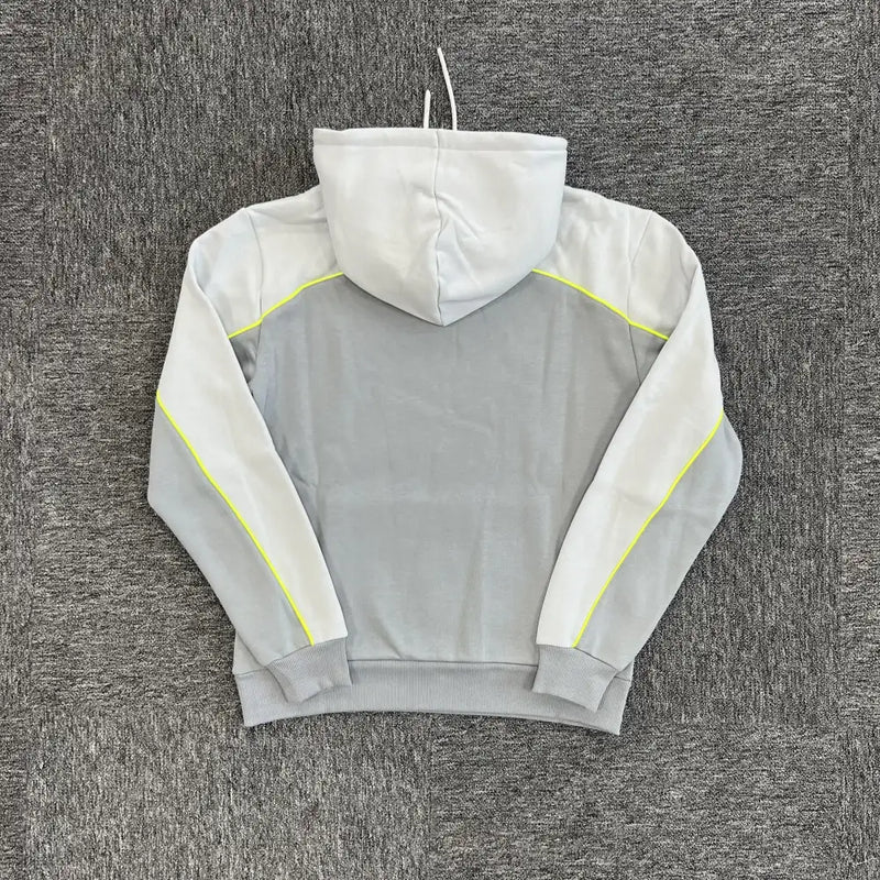 The Syna World Grey Colorblock Neon Trim Hoodie, featuring white sleeves, a white hood, and yellow piping along the sleeves, is laid flat on a textured gray surface and displayed from the back.