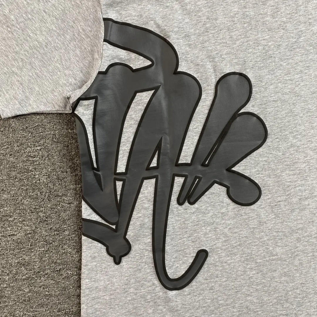 A close-up of two Syna World Grey Letter Graphic Crewneck T-Shirts, both 100% cotton, laid side by side on a textured grey surface, with a bold black abstract design spanning across both shirts.