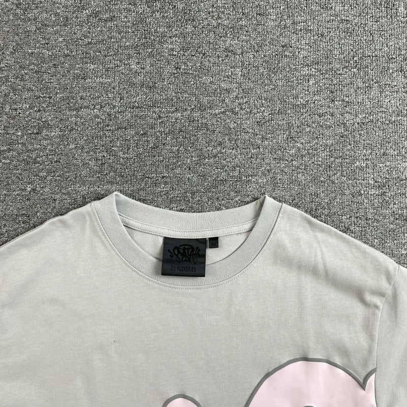 A close-up of the collar and shoulders of the Syna World Grey Pink Letter Print Short Sleeve T-Shirt shows its black label and oversized graphic print with pink accents, displayed on a textured gray surface.