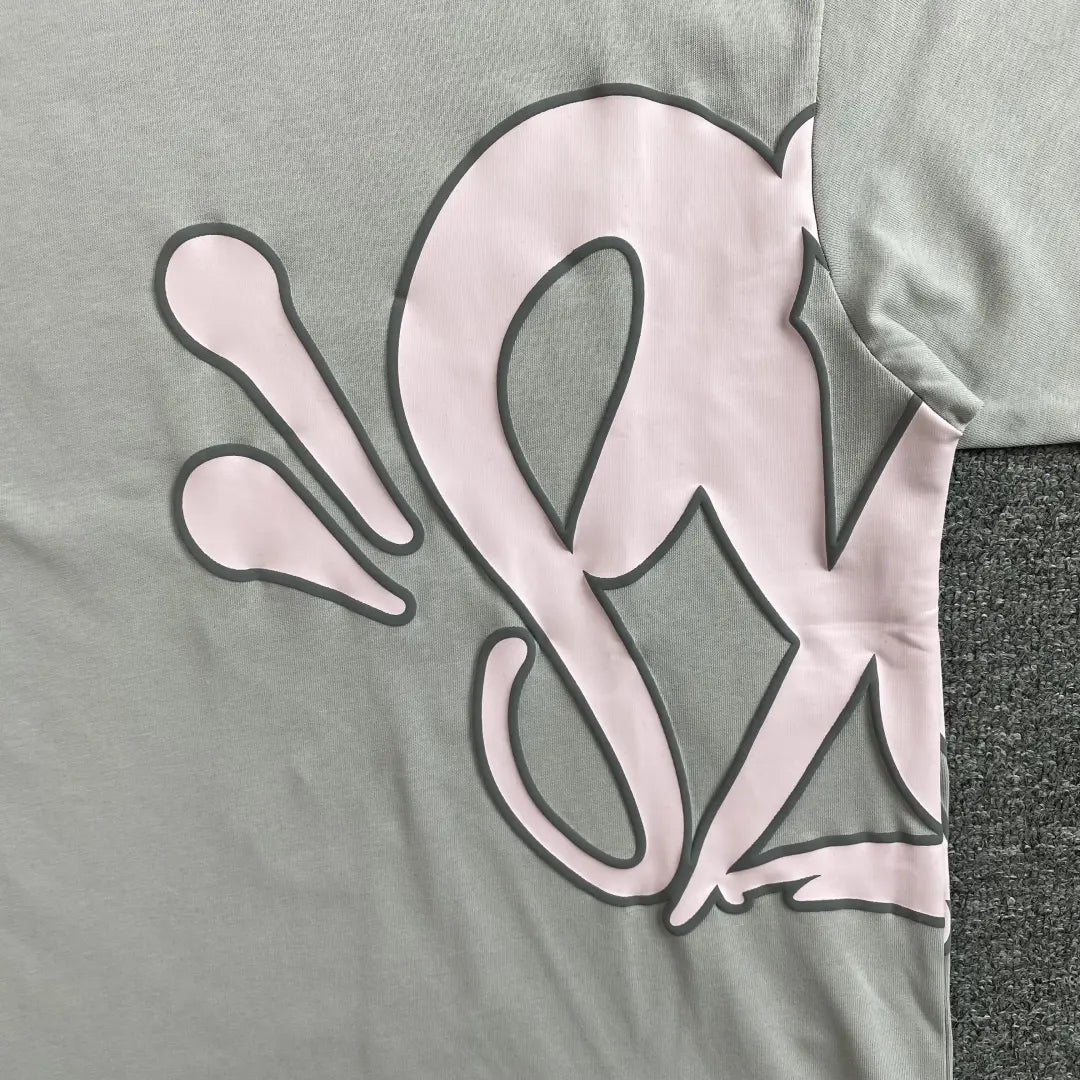 The Syna World Grey Pink Letter Print Short Sleeve T-Shirt features a large pink and gray graphic with curved, abstract shapes on one side and sleeve, displayed flat on a textured surface.