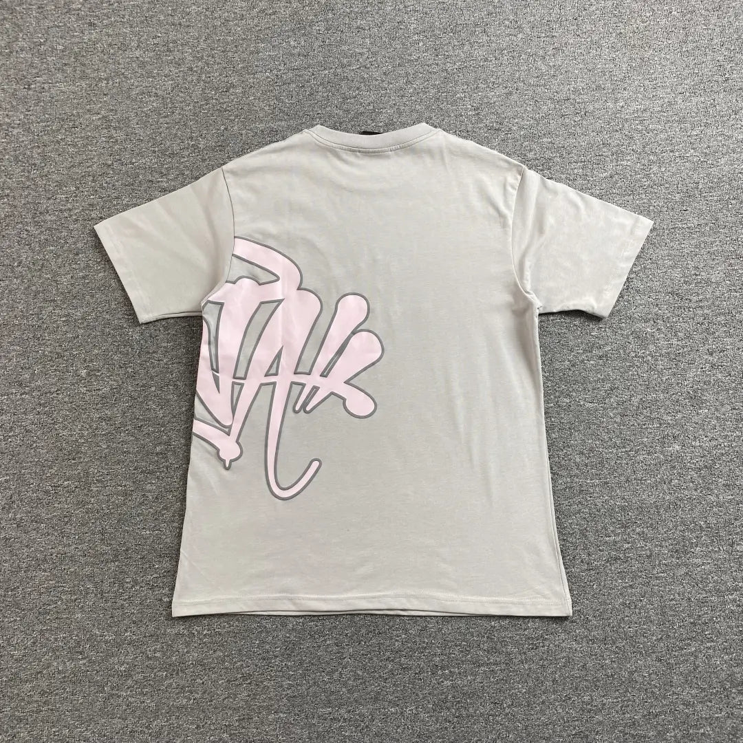 The Syna World Grey Pink Letter Print Short Sleeve T-Shirt, made of cotton, is laid flat on a gray carpet and features an oversized pink graffiti-style graphic on the lower left back—a standout streetwear piece.