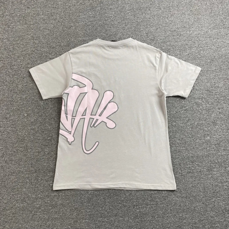 The Syna World Grey Pink Letter Print Short Sleeve T-Shirt, made of cotton, is laid flat on a gray carpet and features an oversized pink graffiti-style graphic on the lower left back—a standout streetwear piece.