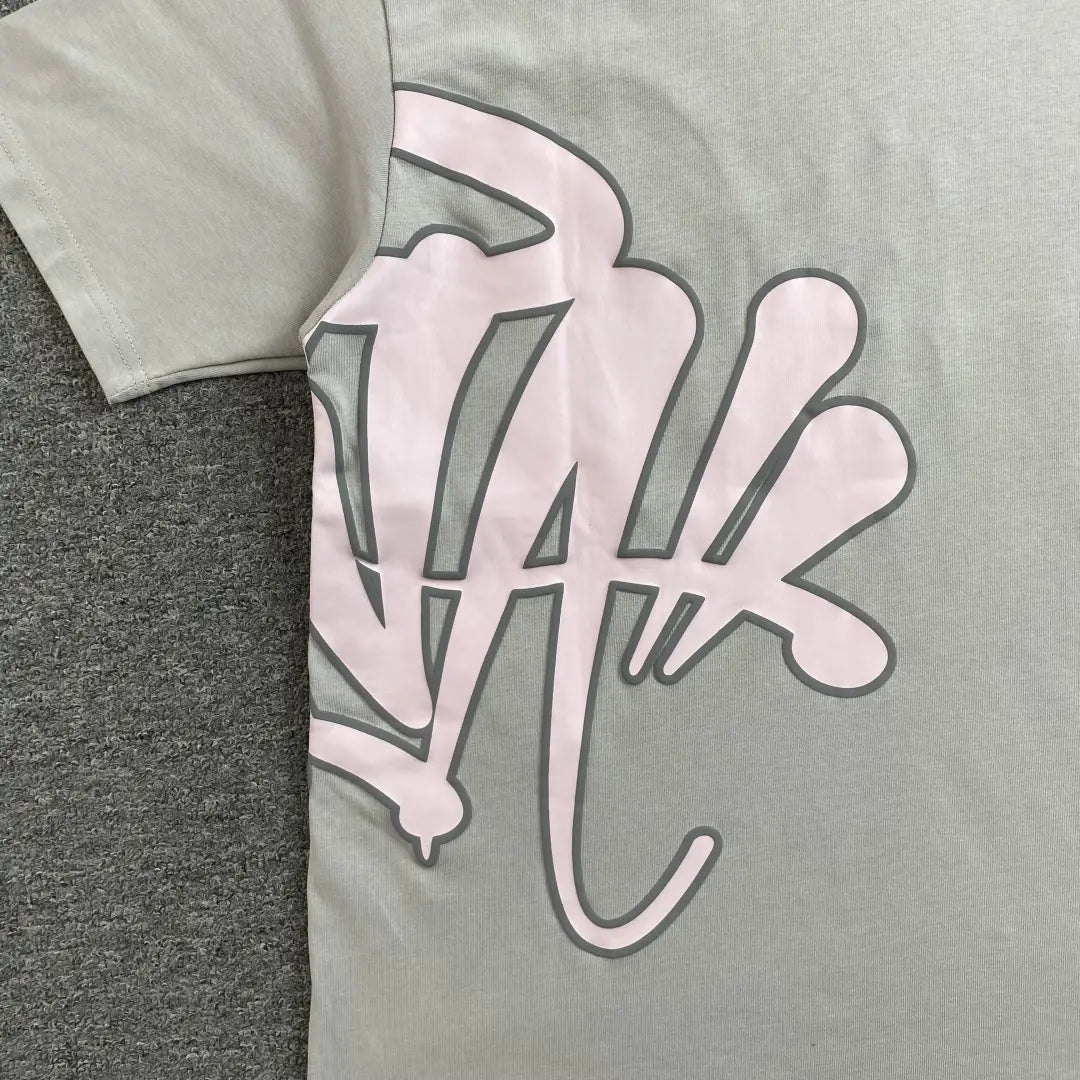 The Syna World Grey Pink Letter Print Short Sleeve T-Shirt, made of light gray cotton, features a bold pink and black graphic on the front and is shown lying on a dark gray textured surface.