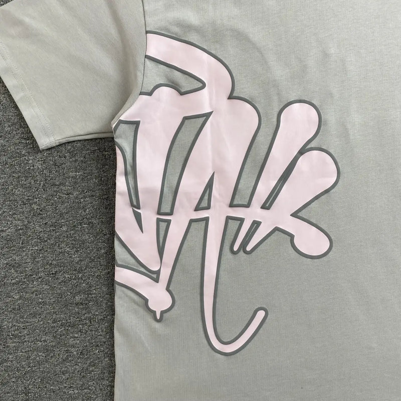 The Syna World Grey Pink Letter Print Short Sleeve T-Shirt, made of light gray cotton, features a bold pink and black graphic on the front and is shown lying on a dark gray textured surface.