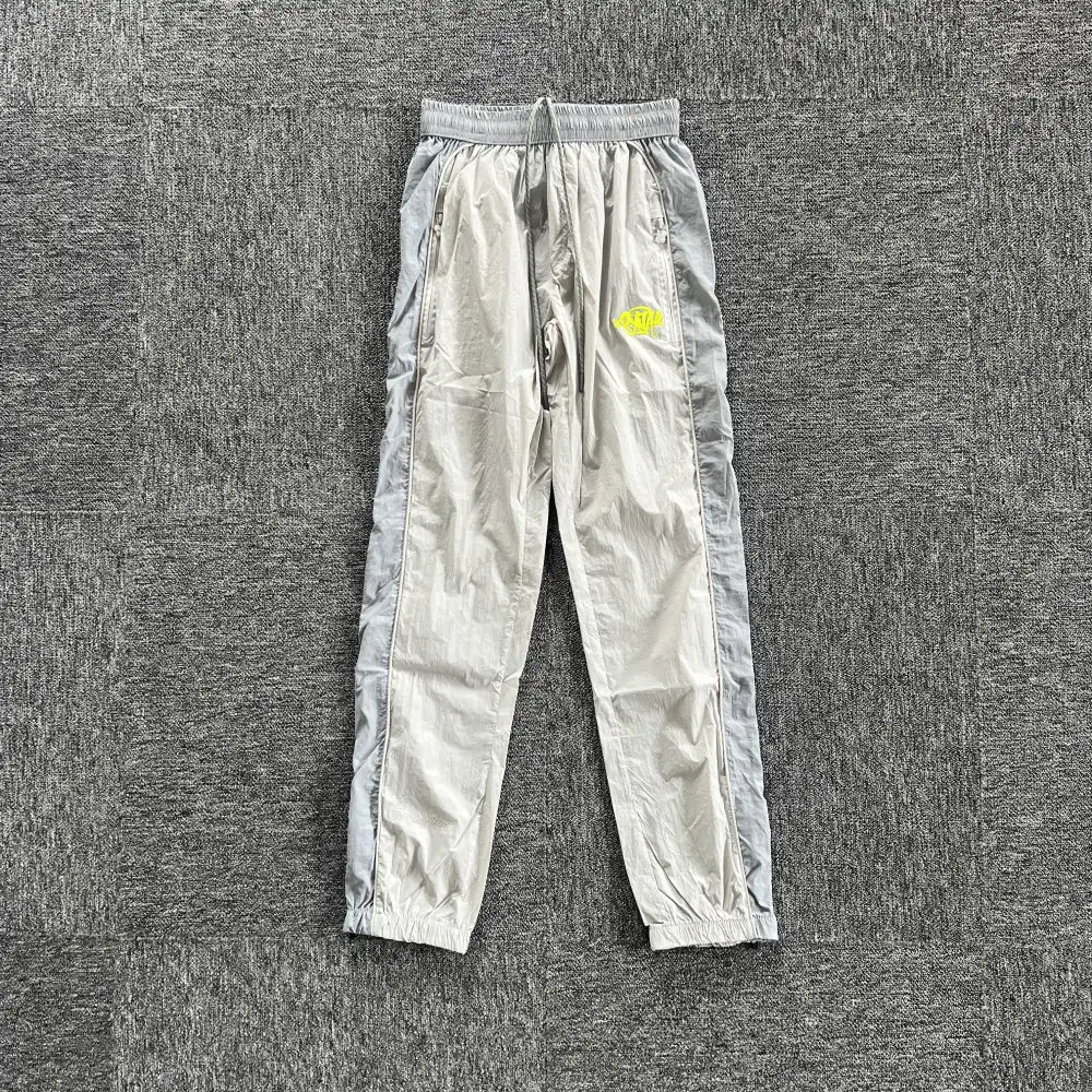 Light grey Syna World track pants with neon yellow embroidered logo on left thigh, laid flat