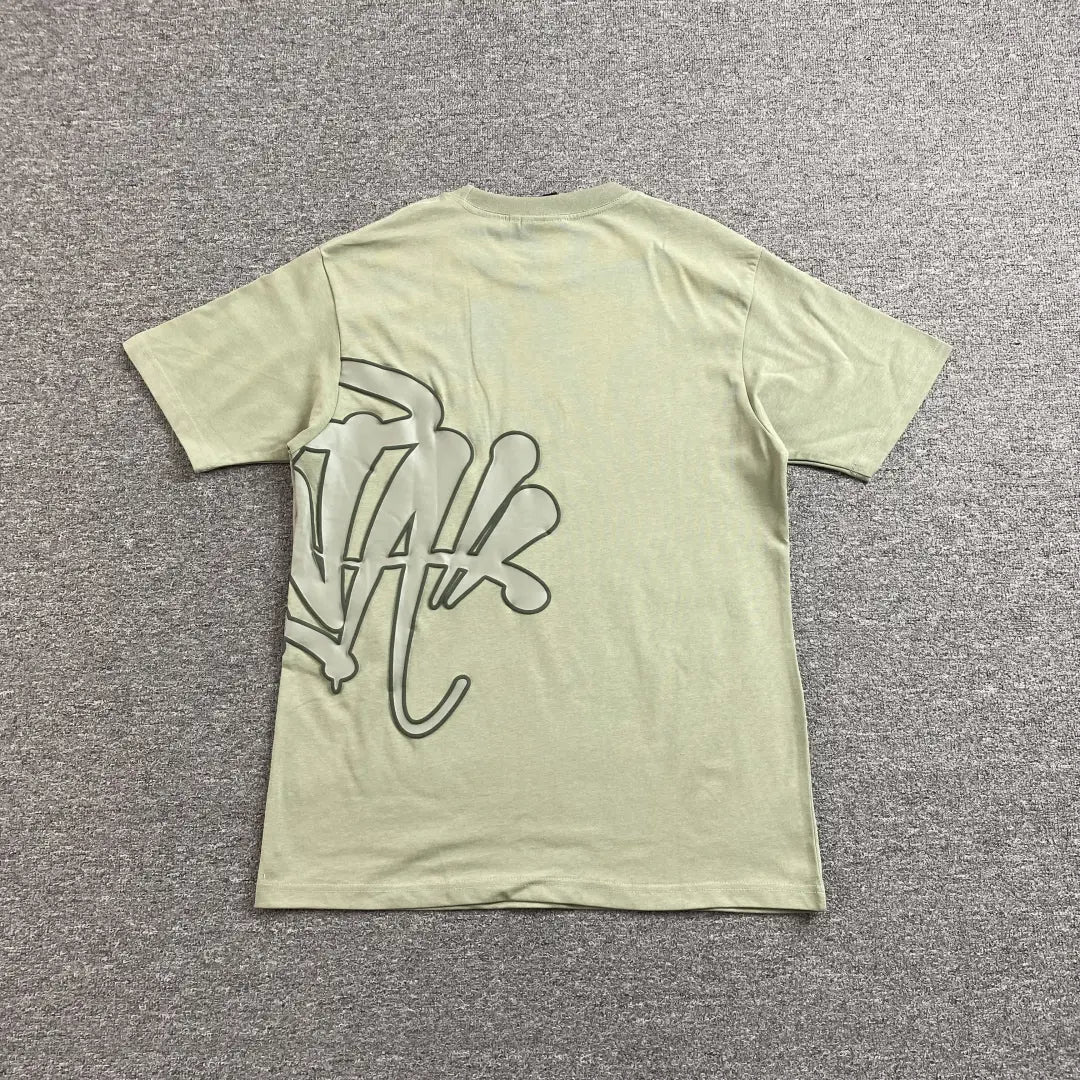 The Syna World Green Letter Graphic Crew Neck T-Shirt lies flat on a gray carpet. This oversized tee has a bold black graphic on the lower left back, making it perfect for adding a streetwear edge to your outfit.