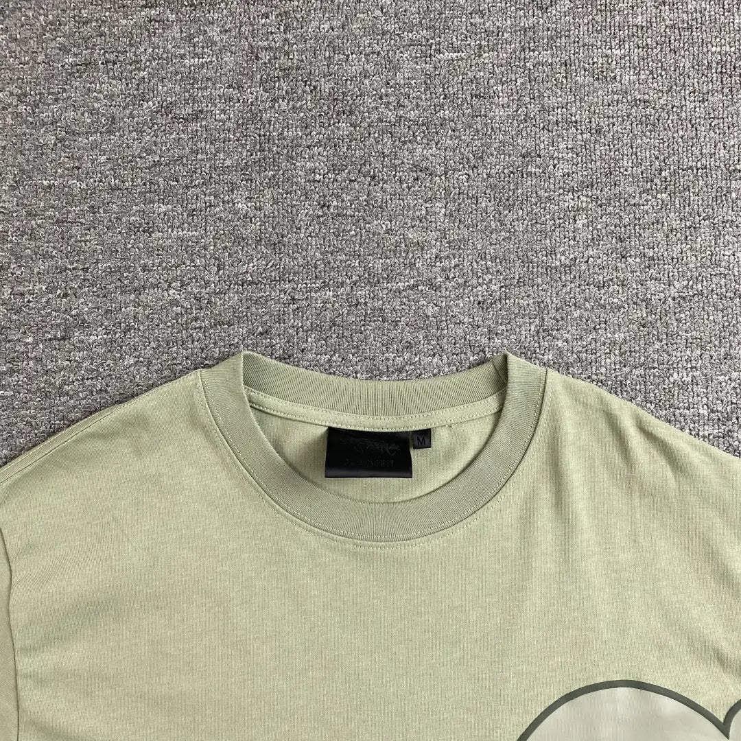 The Syna World Green Letter Graphic Crew Neck T-Shirt, shown flat on a gray textured surface with its black collar tag visible, features a hint of its oversized graphic, making it ideal for laid-back streetwear style.