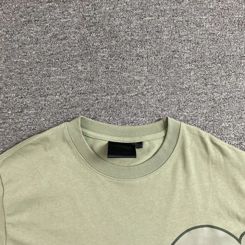 The Syna World Green Letter Graphic Crew Neck T-Shirt, shown flat on a gray textured surface with its black collar tag visible, features a hint of its oversized graphic, making it ideal for laid-back streetwear style.