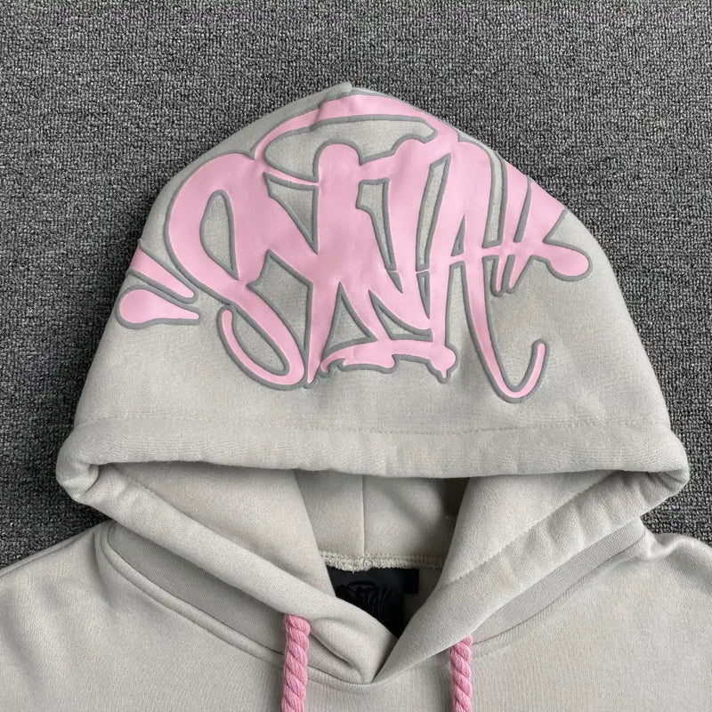 The Syna World Grey Pink Graphic Hood Oversized Hoodie features bold pink graffiti-style lettering on the hood and pink drawstrings, making it a standout streetwear piece in light gray, shown on a textured gray surface.