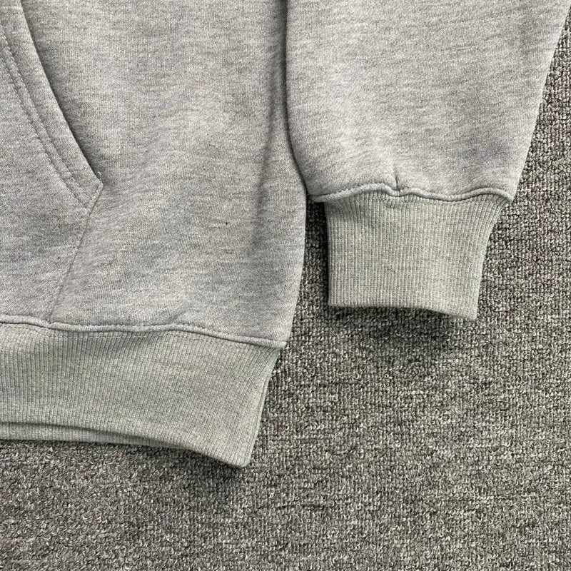 A close-up of the lower section of the Syna World Grey Pink Graphic Hood Oversized Hoodie shows its ribbed cuff and pocket, laid on a textured gray carpet.