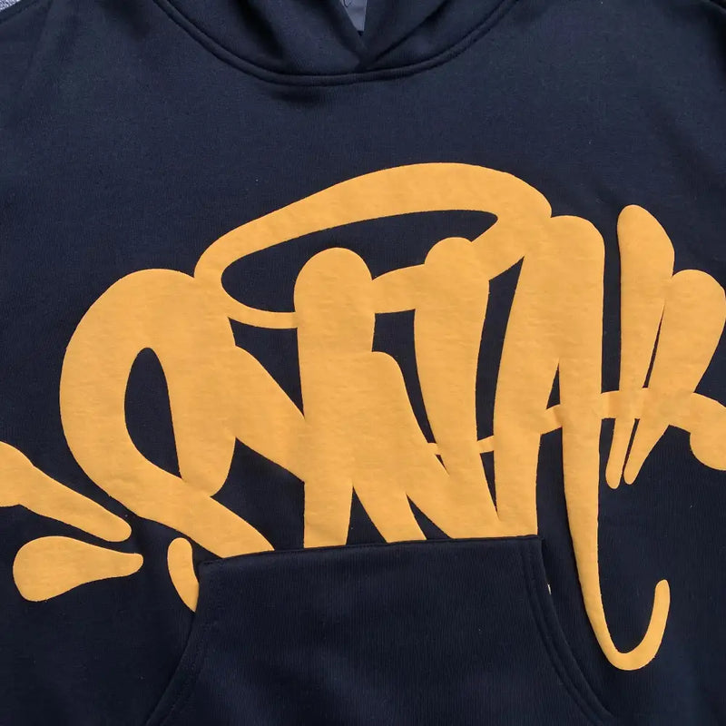 A close-up of the Syna World Black Yellow Graffiti Graphic Oversized Hoodie highlights bold yellow graffiti lettering on the chest and above the pocket—ideal for streetwear pullover fans.