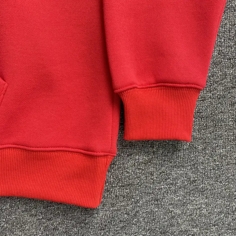 A close-up of the bottom hem and sleeve cuff of the Syna World Red Graffiti Letter Print Oversized Hoodie, featuring ribbed fabric, laid on a textured gray surface.