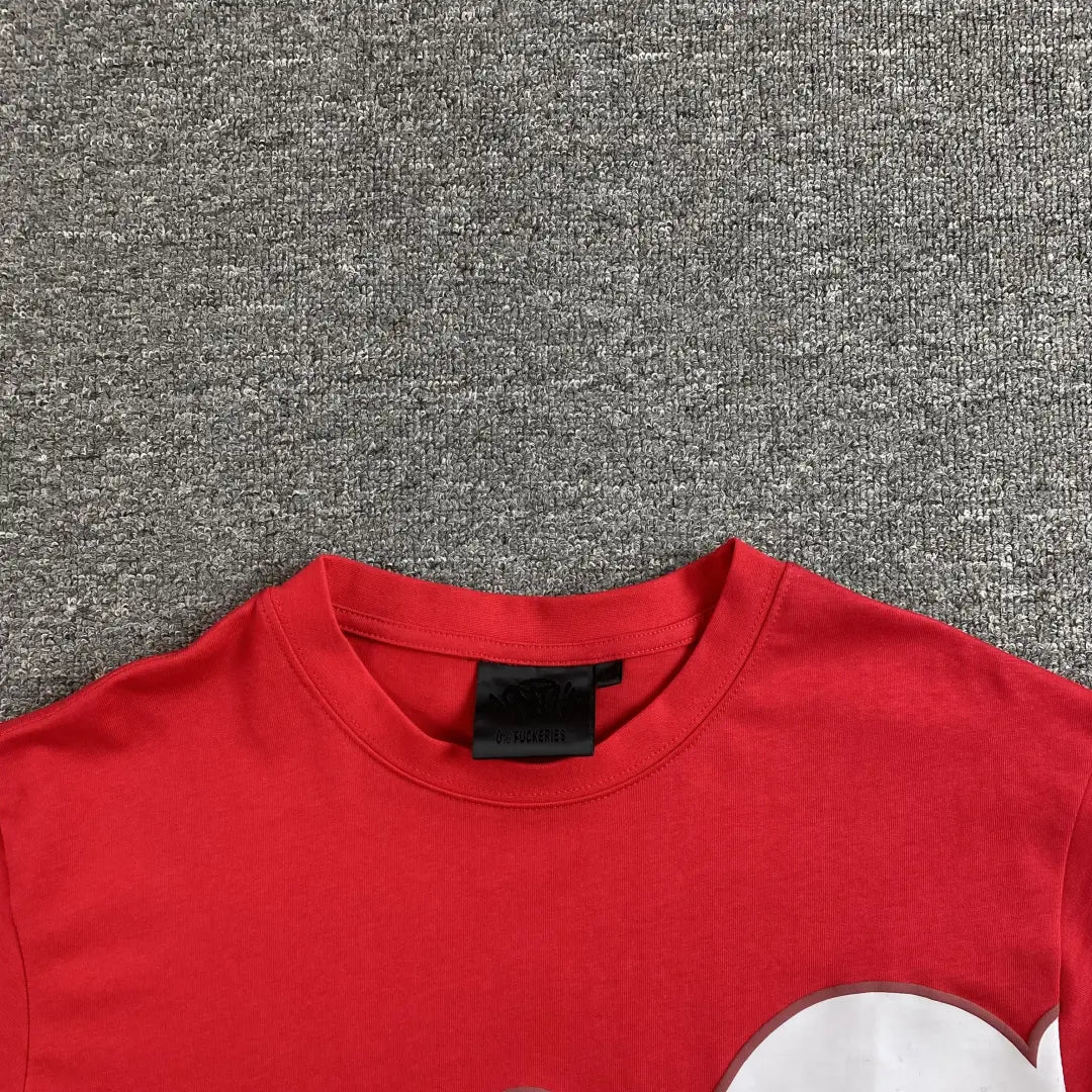 A Syna World Red Letter Print Crew Neck T-Shirt with a black neck tag is displayed on a grey textured surface, showing only the upper part of this streetwear cotton piece.