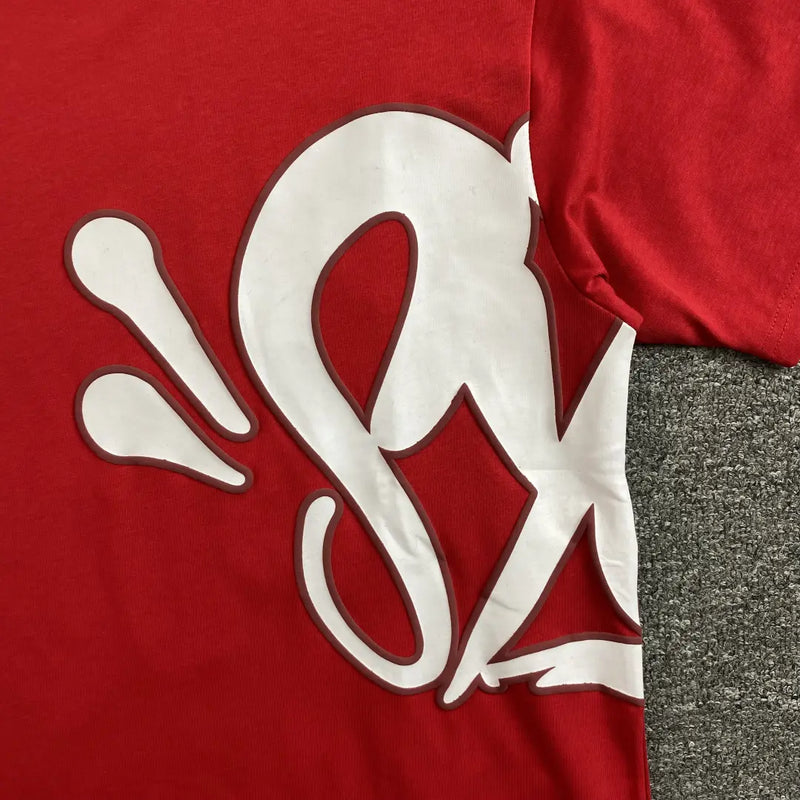 The Syna World Red Letter Print Crew Neck T-Shirt features bold white graphics with curved lines extending from front to side, giving this streetwear cotton tee a unique look. Shown laid flat on a gray textured surface.