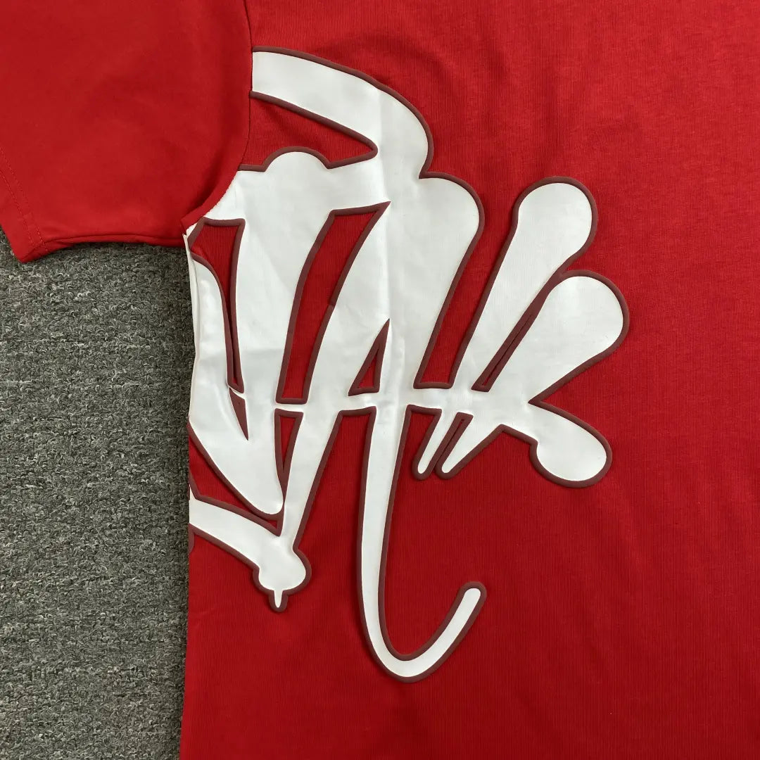The Syna World Red Letter Print Crew Neck T-Shirt lies flat on a gray carpet, showcasing its bold, oversized white graffiti-style print that stretches from the side seam across the front.
